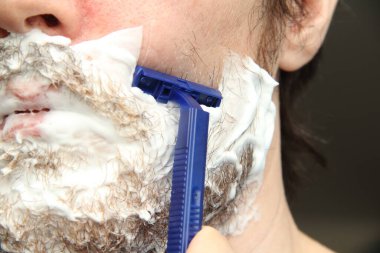 Bir adam mavi tek kullanımlık tıraş bıçağıyla sakalını kesiyor. Yanaklarında ve çenelerinde beyaz tıraş köpüğü.