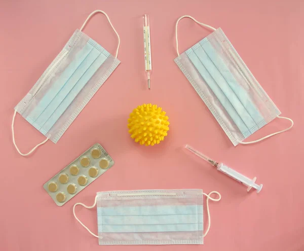 Medical disposable masks, thermometer, syringe and pills on a pink background surround the coronovirus in the form of a yellow ball with spikes. Coronovirus Outbreak Defense Concept