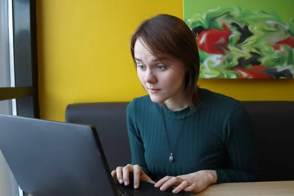 Menina bonita sentada em um café em uma mesa com um laptop. Funciona no computador e digita no teclado. Olha para o ecrã com uma expressão emocional. No rosto é uma expressão de — Fotografia de Stock
