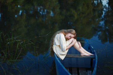 Batık bir teknede oturan uzun saçlı sarışın, melankolik, depresyonlu.