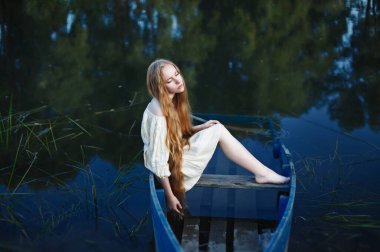 Batık bir teknede oturan uzun saçlı sarışın, melankolik, depresyonlu.
