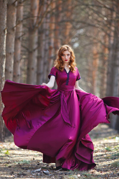 red-haired girl in a burgundy dress walks in the forest