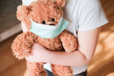Koronavirüsten korumak için tıbbi maskeli oyuncak ayı tutan küçük bir kız. Evde konsept kal. Virüs salgını önleme.