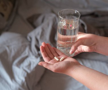 Elinde haplar ve bir bardak su tutan ve yatağın yanında duran genç bir kadının elleri..