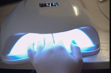 Kadın ellerinin yeni, güzel, modern jöle cilası manikürünü yaptığı yakın çekim görüntüsü. Kadın, çiviyi kurutmak için ultraviyole lambanın içine elini sokuyor. Yatay görüş. Coronavirus karantinası sırasında ev manikürü.