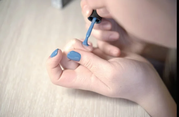 Young woman applying nail polish with brush from bottle, polishing painting fingernails with blue color enamel, doing manicure at home, perfect healthy nails care