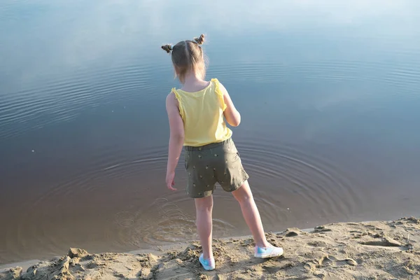 little cute girl throwing stones to water. girl with space buns on sandy beach playing with stones. back view. summer holiday and relax time for children.