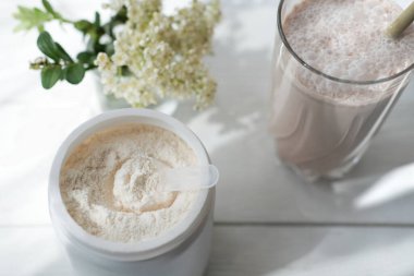 Büyük bardakta beyaz şarap. beyaz protein çikolatalı kokteyl. Protein tozu dolu bir kavanoz ve beyaz bir masanın üzerinde çiçekler. Sağlık ve güzellik için sabah içkisi. en üstteki görünüm