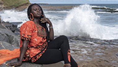 Gana 'lı kadın Takoradi' de deniz kenarındaki kayalıklarda turuncu Afrika elbisesi giyerek oturuyor..
