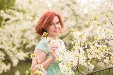 Mavi elbiseli güzel kızıl saçlı kadın çiçek açmış elma ağacının altında poz veriyor. 