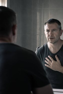 Gri tişörtlü bir adam banyo aynasında kendine bakıyor.