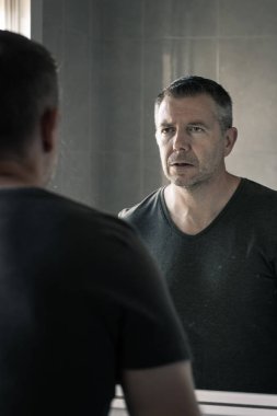 Gri tişörtlü bir adam banyo aynasında kendine bakıyor.