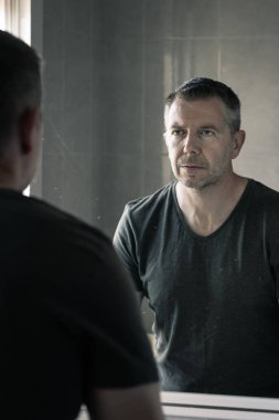 Gri tişörtlü bir adam banyo aynasında kendine bakıyor.