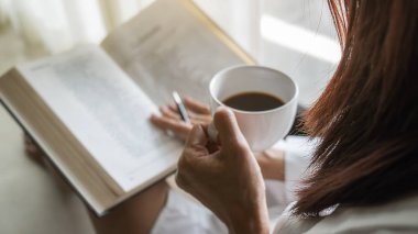 Asyalı kadın elinde bir fincan kahve tutuyor ve sabah kalktıktan sonra pencerenin yanında kitap okuyor. Sabah yaşam tarzı konsepti.