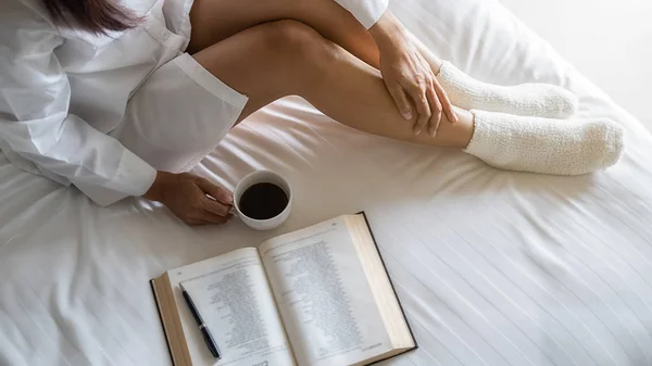 Ázsiai nő kezében egy csésze kávét, és olvasni egy könyvet az ágyon után kelni reggel. Reggeli életmód koncepció. — Stock Fotó