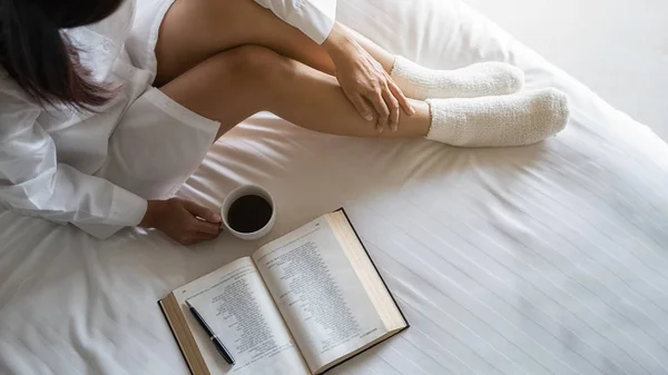 Ázsiai nő kezében egy csésze kávét, és olvasni egy könyvet az ágyon után kelni reggel. Reggeli életmód koncepció. — Stock Fotó