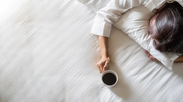 Itt az ideje kávézni. Ázsiai lusta nő fekszik, és egy csésze kávét tart az ágyon reggel, lágy reggeli fényben. Reggeli életmód koncepció. — Stock Fotó