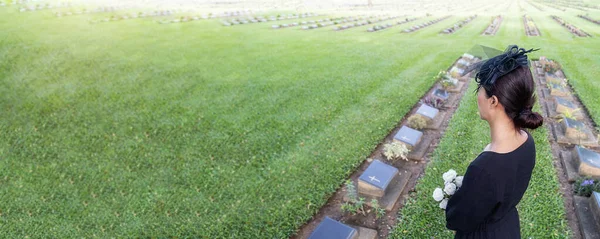 Mourning young woman holding white flowers at her family grave in beautiful green cemetery.