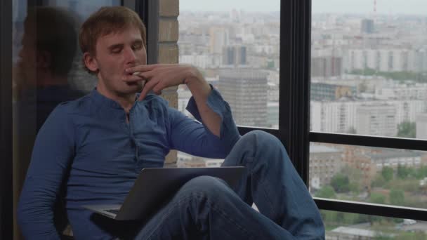 Чоловік з ноутбуком сидить на підвіконні і дивиться у вікно хмарочоса — стокове відео