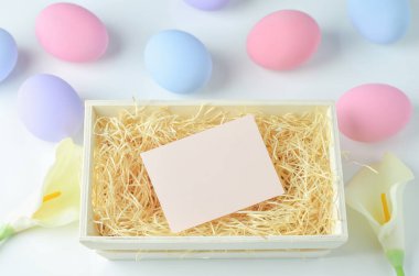 Ahşap sepetteki boş tebrik kartı beyaz masa arkasında ahşap yün pastel ester yumurtaları ve kalla zambağı çiçeği.