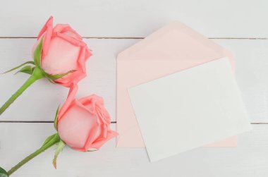 İçinde pembe zarf ve pembe gül çiçekleri olan boş bir tebrik kartı beyaz ahşap arka planda yumuşak bir tonda, Sevgililer Günü gülü ve kartı.