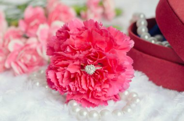 Pembe karanfil çiçeğiyle nişan yüzüğü inci kolye ve beyaz kürk arka planda kırmızı mücevher kutusu.