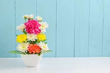 Beyaz vazoda bir demet renkli çiçek, pembe karanfil ve kasımpatı çiçekleri mavi ve beyaz ahşap arka planda