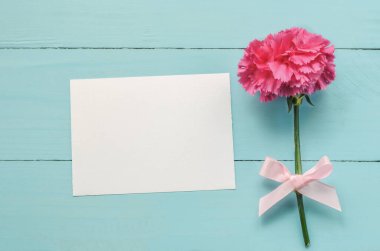 İçinde pembe karanfil çiçeği ve mavi ahşap arka planda kurdele olan boş beyaz tebrik kartı.