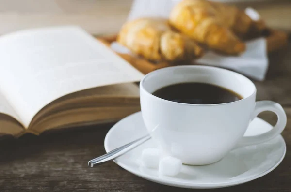 Close up of black coffee with book and croissant on wooden table with vintage and vignette tone