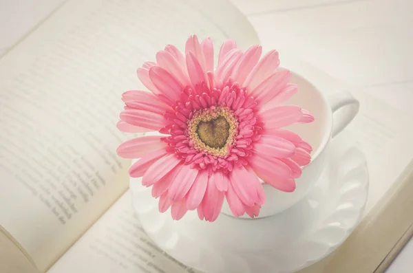 Close up of pink Gerbera daisy flower and white coffee cup on opened book with soft vintage tone