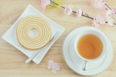 Baumkuchen tatlı katmanlı kek beyaz tabakta bir fincan sıcak çay ve yapay pembe sakura çiçeğiyle ahşap masa üzerinde klasik tonda.