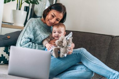 Güzel anne bebeğiyle vakit geçirirken dizüstü bilgisayar kullanıyor ve gülümsüyor..