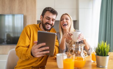 Genç bir çift modern ışık mutfağında kahvaltı yapıyor. Mutlu çift tablet kullanıyor ve birlikte kahvaltı yapıyor..