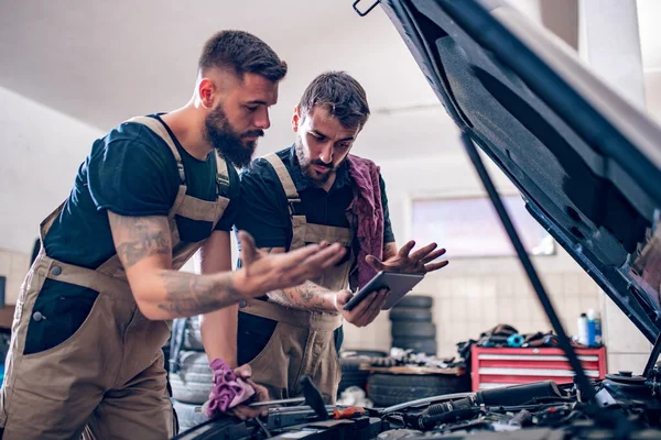 Two mechanics working on reports in a workshop.Mechanics with scan tool diagnosing car in open hood.