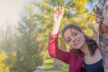 Palmiye ağacının arkasından bakan ve elinde bardaklarla el sallayan komik bir genç kadın.