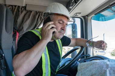 Kasklı ve güvenlik gözlüklü bir kamyon şoförü telefonla konuşuyor ve haritaya bakıyor.