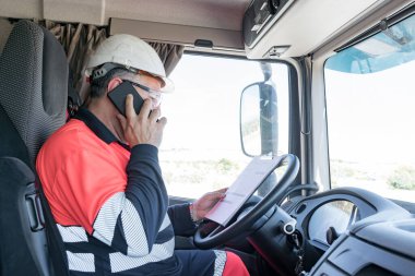 Kasklı, güvenlik gözlüklü ve yüksek görüş mesafesi olan bir kamyon şoförü telefonla konuşuyor.