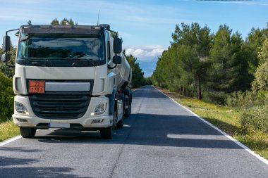 Fuel tank truck driving on a narrow, straight road lined with trees clipart