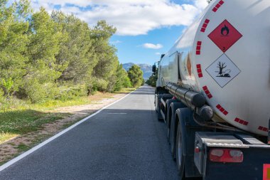 Dar, ağaçlarla kaplı düz bir yolda giden bir yakıt tankında yanıcı ve kirletici sıvılar için tehlikeli etiketler..