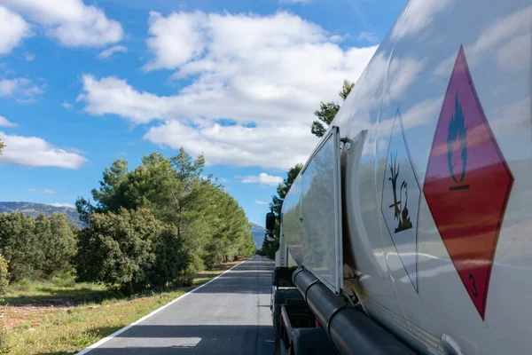 Dar, ağaçlarla kaplı düz bir yolda giden bir yakıt tankında yanıcı ve kirletici sıvılar için tehlikeli etiketler..
