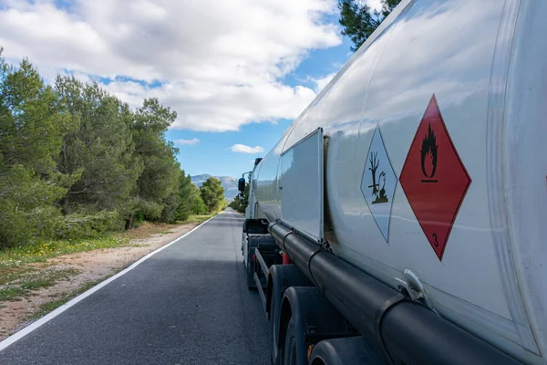 Dar, ağaçlarla kaplı düz bir yolda giden bir yakıt tankında yanıcı ve kirletici sıvılar için tehlikeli etiketler..