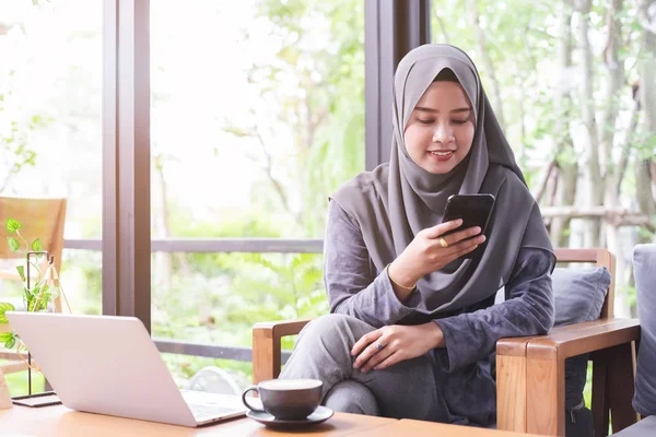 Asiático jovem muçulmano mulher sentado no café manhã com laptop na mesa e usando smartphone . — Fotografia de Stock