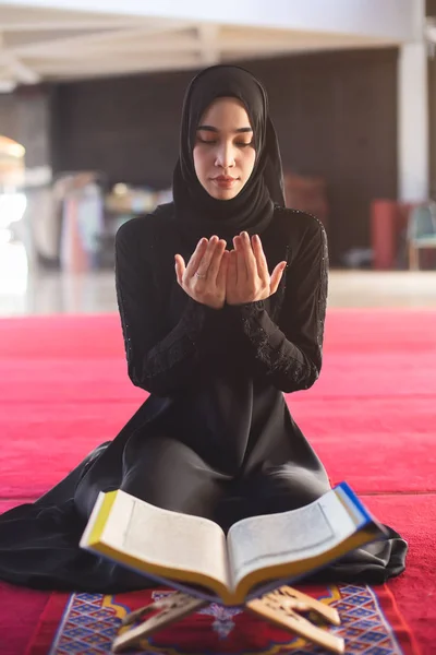 Jonge moslimvrouw in zwarte jurk biddend met koran in moskee. — Stockfoto