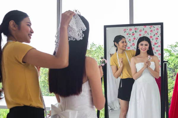 Reflection in mirror of asian bride trying on wedding dress in shop with women assistant.