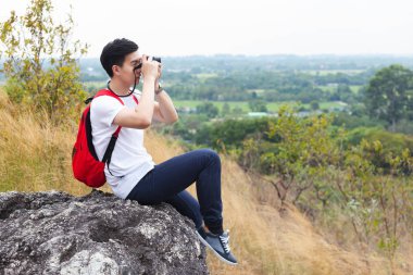 Sırt çantalı genç turist kayalık dağda oturuyor ve kameradan fotoğraf çekiyor. Seyahat kavramı.