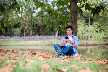 Parkta ağacın altında akıllı telefon kullanan yakışıklı adam..