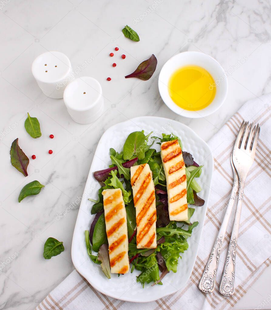 Green salad with fried halloumi cheese in a white plate on a marble background, top view. 