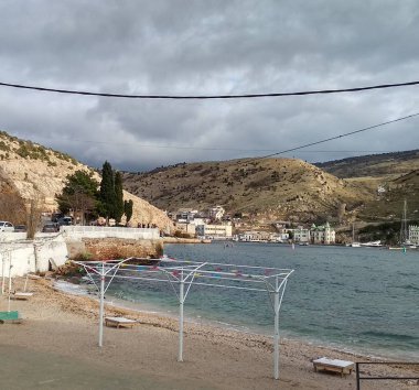 Kışın Balaklava Körfezi 'nde kumsal. Boş güneşlik. Karanlık su. Kurşun bulutlar. Köy. Güçlendirilmiş sahil. Spreyler.