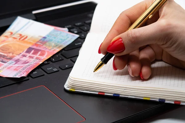 A female hand holds a pen, Swiss banknotes and a notebook lie on the keyboard, the work of a copywriter, reliable earnings during the quarantine period