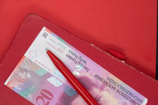 20 frank İsviçre banknotu, eski defter ve bir dizüstü bilgisayar kapağı, hepsi kırmızı renkli.